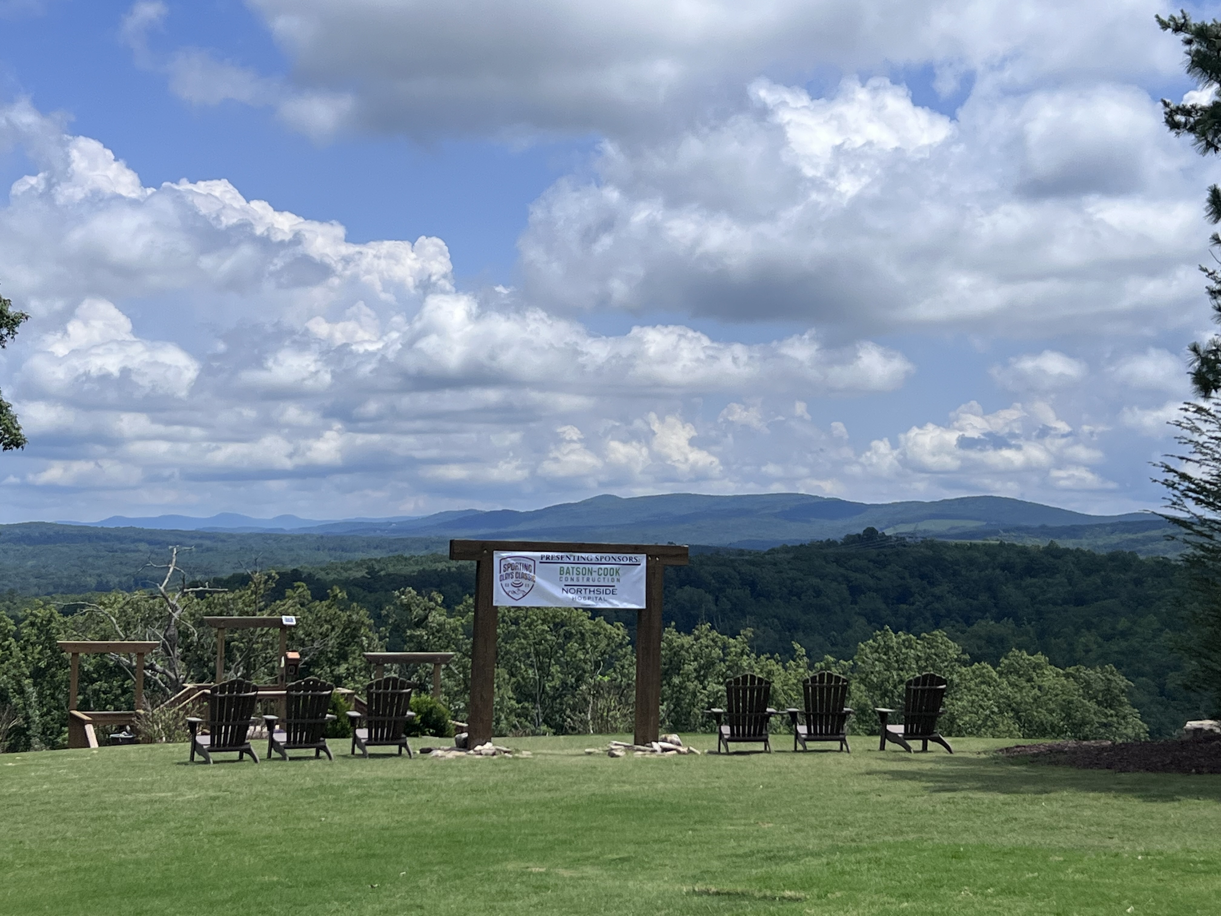 Outdoor seating area with Adirondack chairs on a grassy lawn overlooking green forest and distant mountains, under a partly cloudy sky, with a wooden arch displaying a banner
